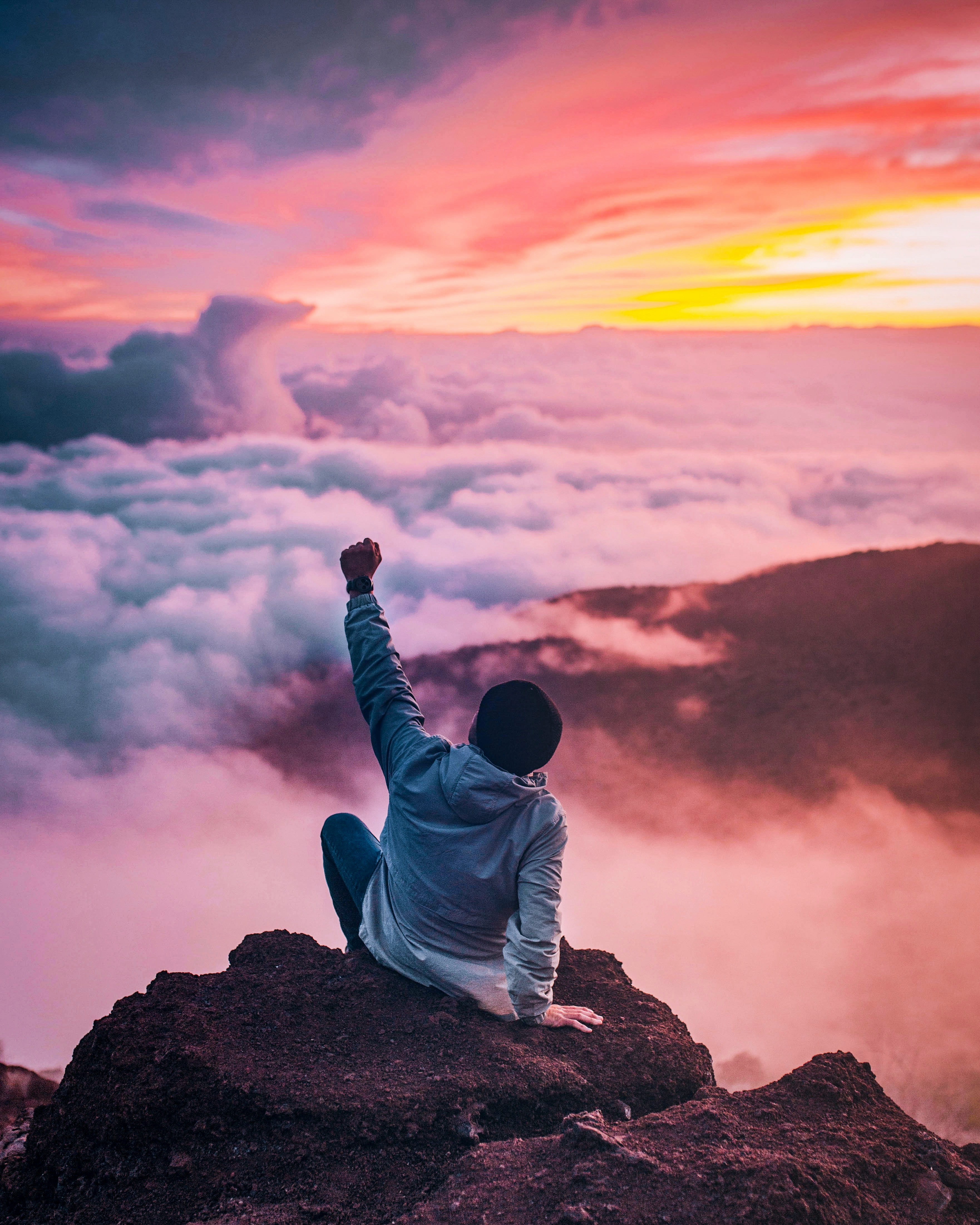 A man standing triumphantly on a mountain peak, raising his fist in the air as he gazes out at expansive clouds and the horizon, symbolizing limitless potential, personal growth, and the power of mindset coaching.